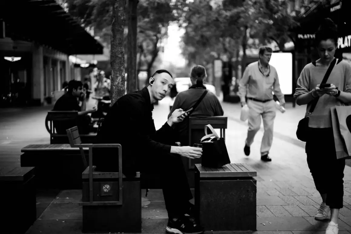 A man with earbuds looks at his phone while sitting on a bench. This 35mm vs 50mm street photography captures a moment of urban life.