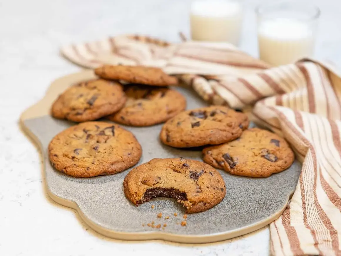 Nybakade chocolate chip cookies recept på en grå platta, med en bit tagen ur en cookie. Två glas mjölk och en randig handduk i bakgrunden.