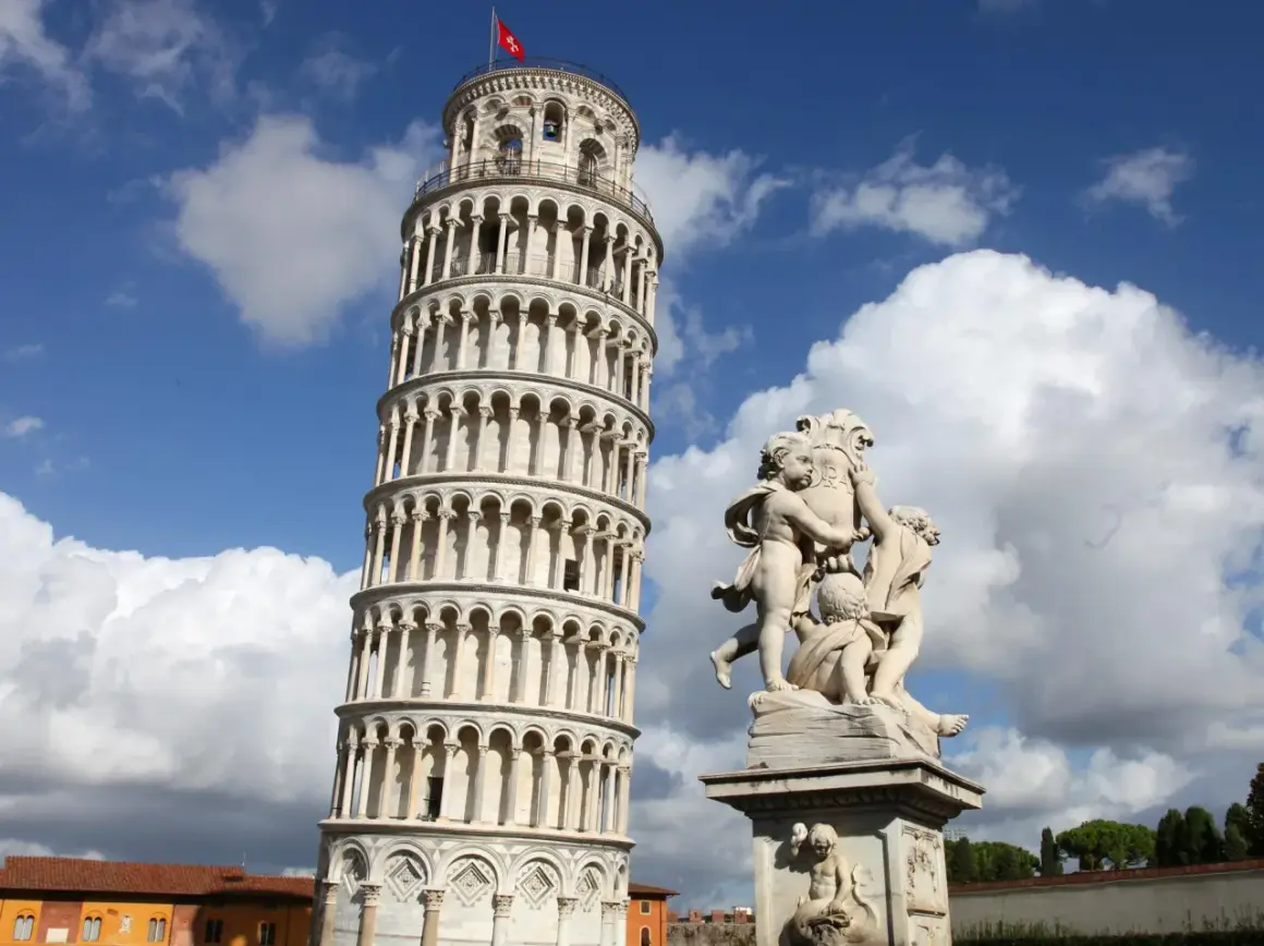 La Torre Pendente di Pisa, con la sua inconfondibile inclinazione, si erge maestosa sotto un cielo azzurro e nuvoloso, accanto a una scultura di putti.