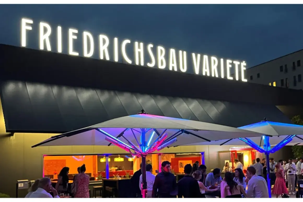 Leuchtende Buchstaben über dem Eingang des Varieté Friedrichsbau. Menschen versammeln sich unter beleuchteten Sonnenschirmen.