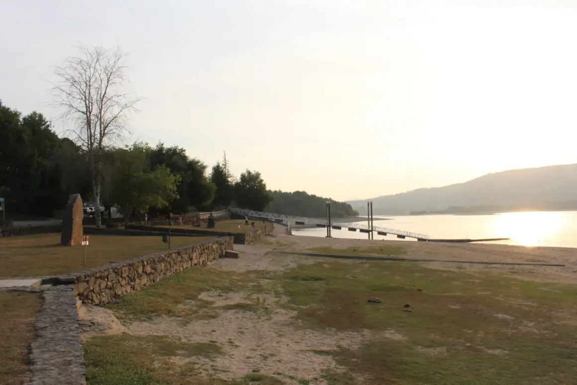 Un tranquilo atardecer en la playa fluvial o corgo, con un muelle y vegetación verde.