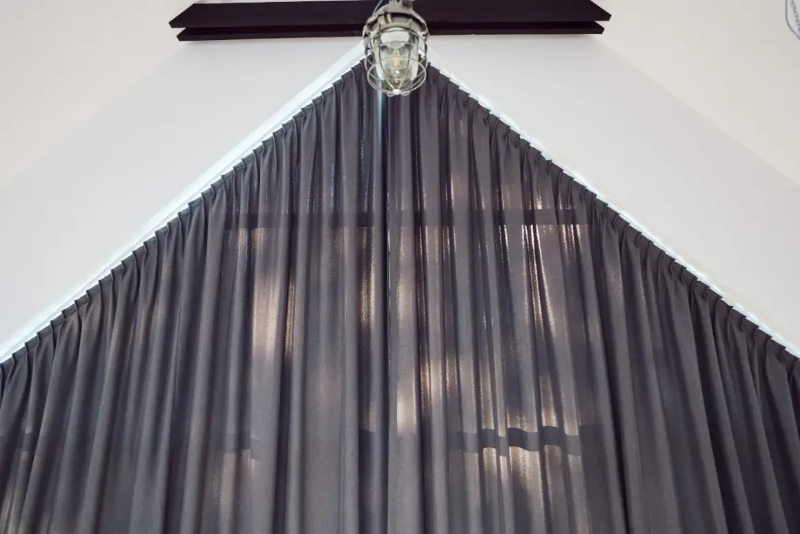 Szare zasłony z połyskującą nitką, idealnie dopasowane do nietypowego okna trapezowego. Nad nimi industrialna lampa.