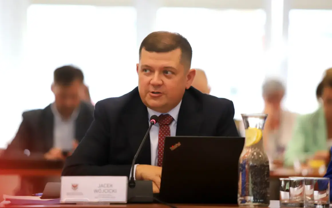 Jacek Wójcicki, siedząc przy stole, przemawia. Jego wzrost jest przeciętny, a na stole leży laptop i woda z cytryną.