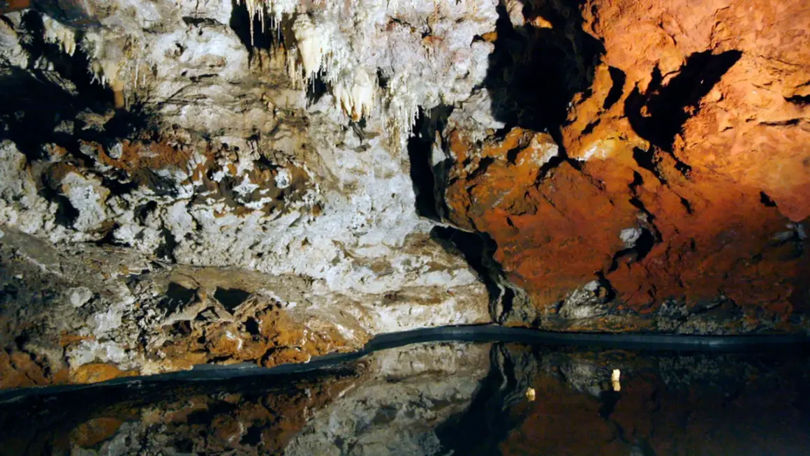 Interior de una cueva con estalactitas y formaciones rocosas rojizas, reflejándose en un espejo de agua. El **cerro del hierro** se revela en su esplendor subterráneo.