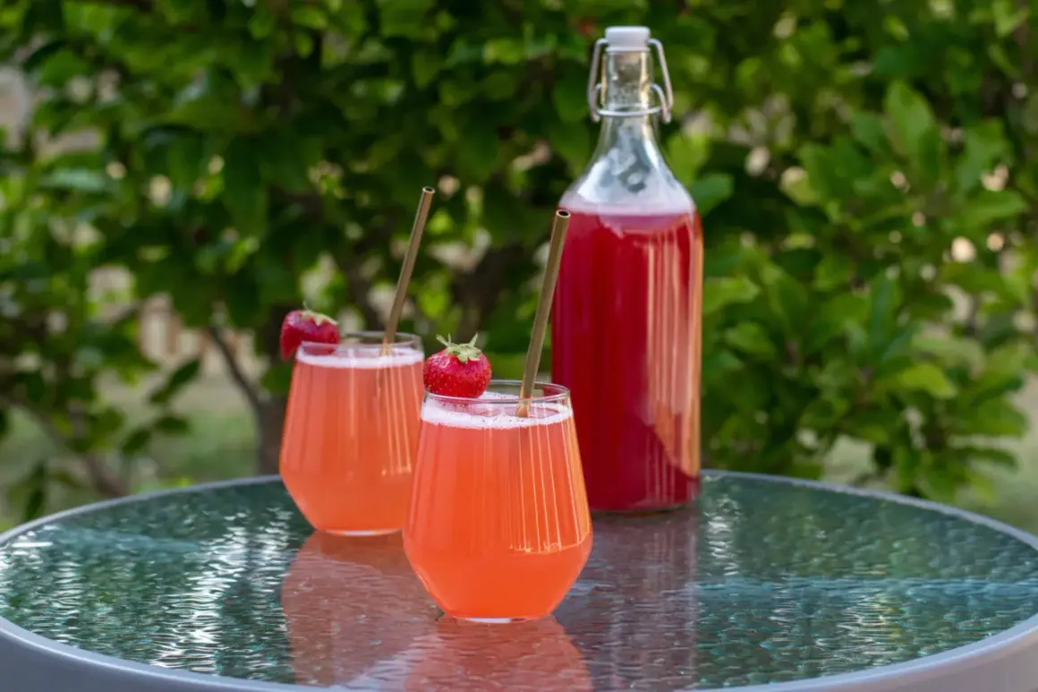 Två glas med rosa rabarbersaft, garnerade med jordgubbar och sugrör, står på ett glasbord. En flaska med saft i bakgrunden. Perfekt för ett rabarbersaft recept.