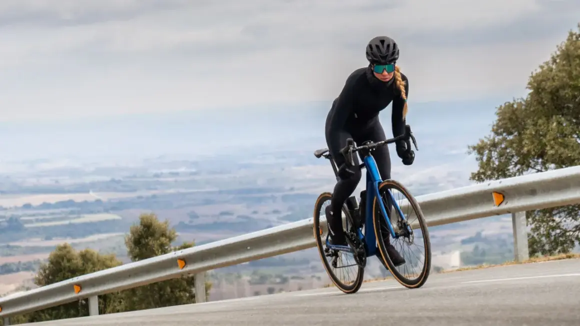 Kobieta w kasku i okularach jedzie na rowerze z niebieskim, aerodynamicznym nadwoziem. Silnik umieszczony centralnie zapewnia moc podczas podjazdu.