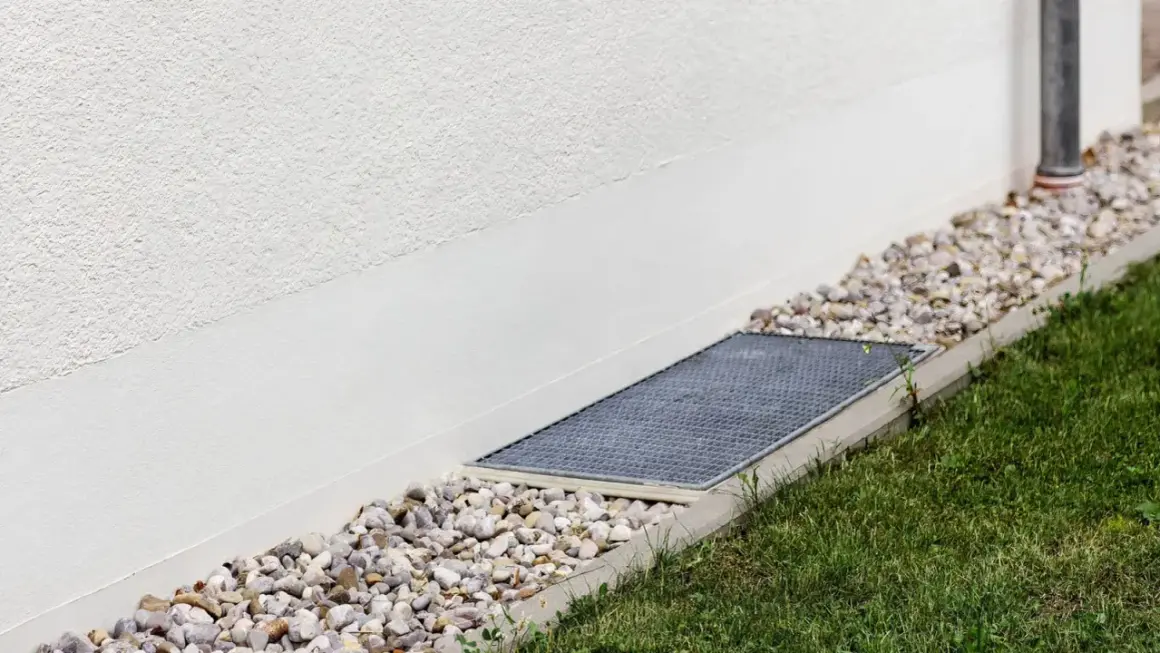 A gravel bed and grate next to a white wall, showing how to level ground for drainage.