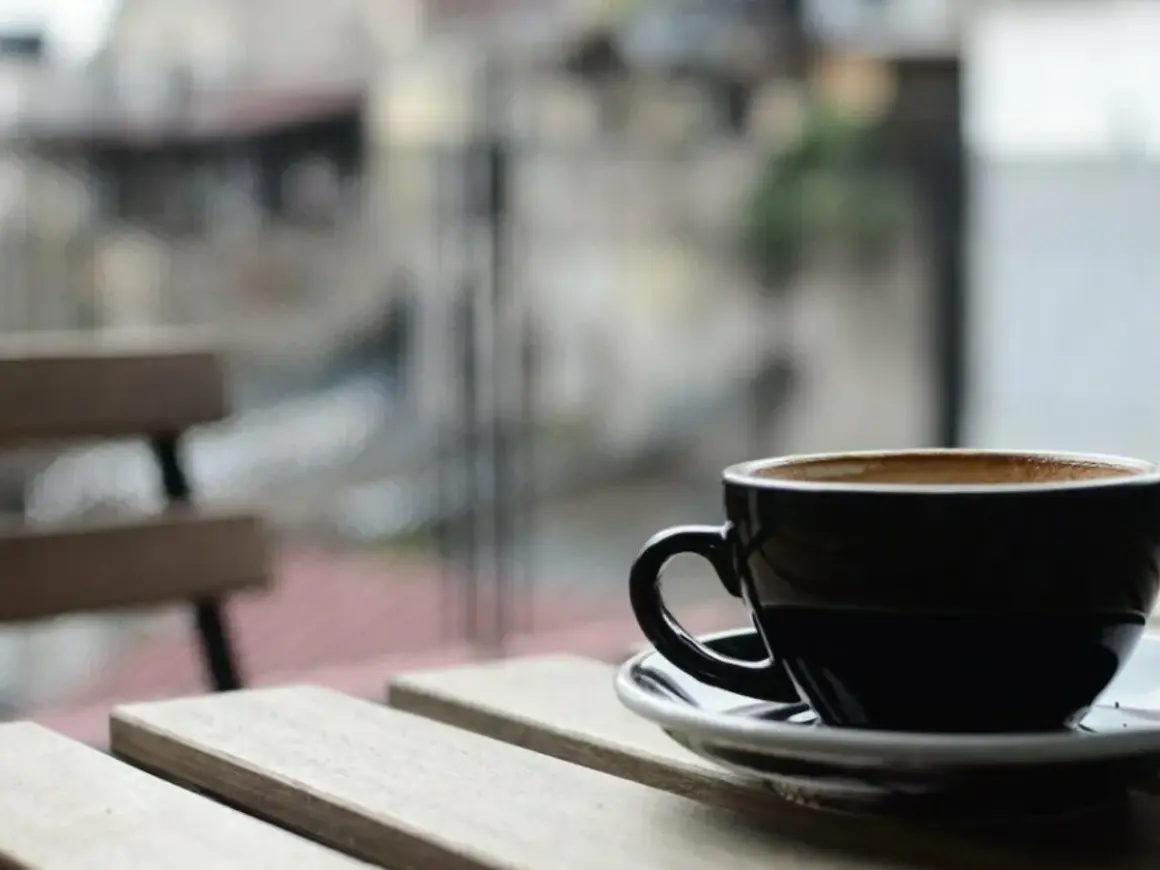 En svart kopp med lungo kaffe står på ett träbord. I bakgrunden syns en bänk och en suddig stadsmiljö.