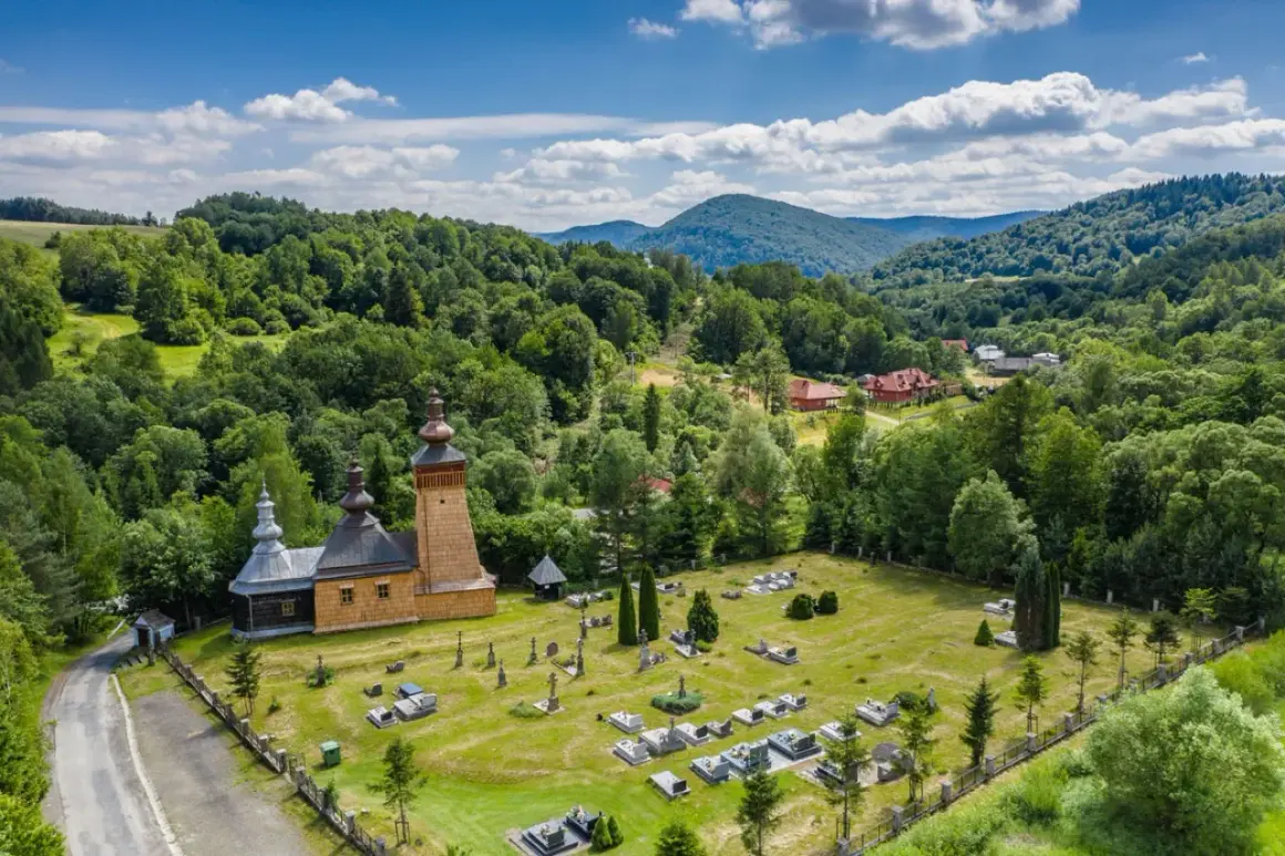 Drewniana cerkiew i cmentarz w malowniczej dolinie Beskidu Niskiego, otoczone bujną zielenią lasów i wzgórz.