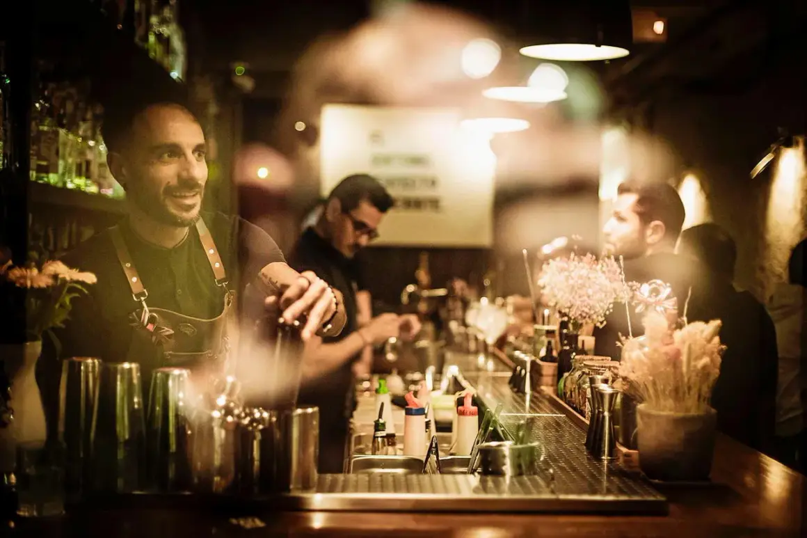 Barman sorridente prepara cocktail dietro un bancone illuminato, perfetto per sapere dove bere a Barcellona.