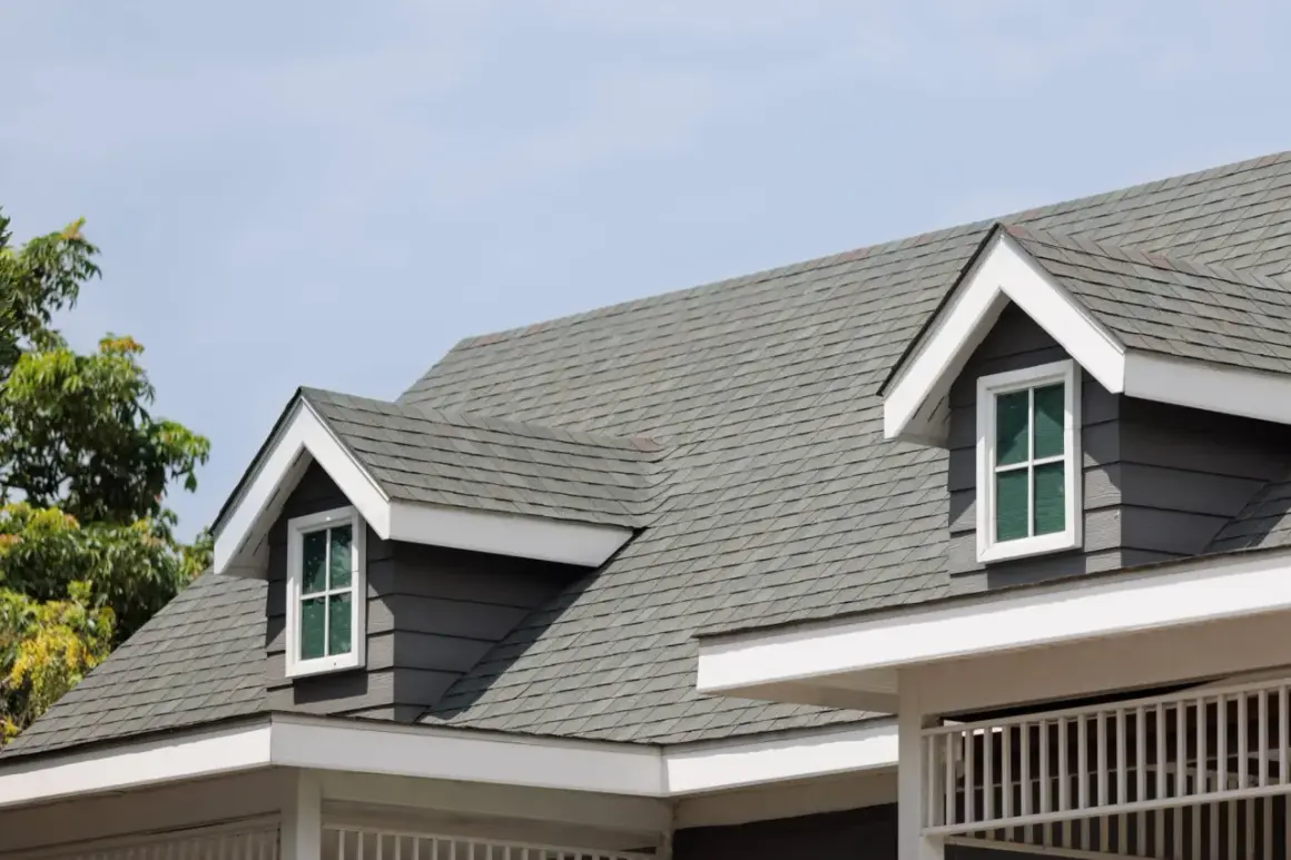 A close-up of a house roof showcasing different types of shingles in a grey hue, with dormer windows peeking out.