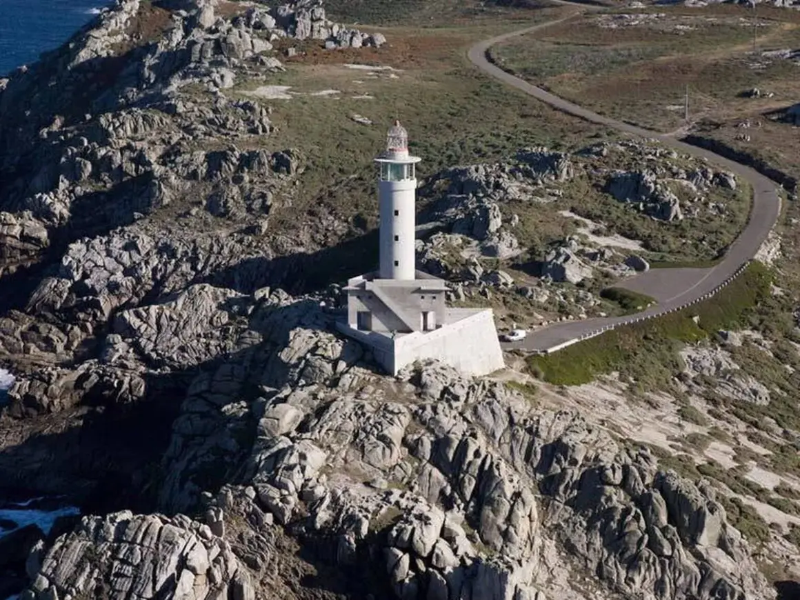 Faro blanco en una punta rocosa, con una carretera serpenteante que sube hacia él.