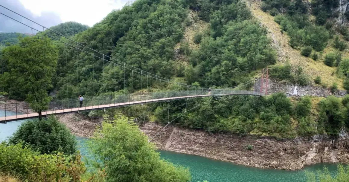Un ponte sospeso attraversa un lago turchese, circondato da montagne boscose. Due persone passeggiano sul ponte, godendosi il panorama dei vagli di sotto.