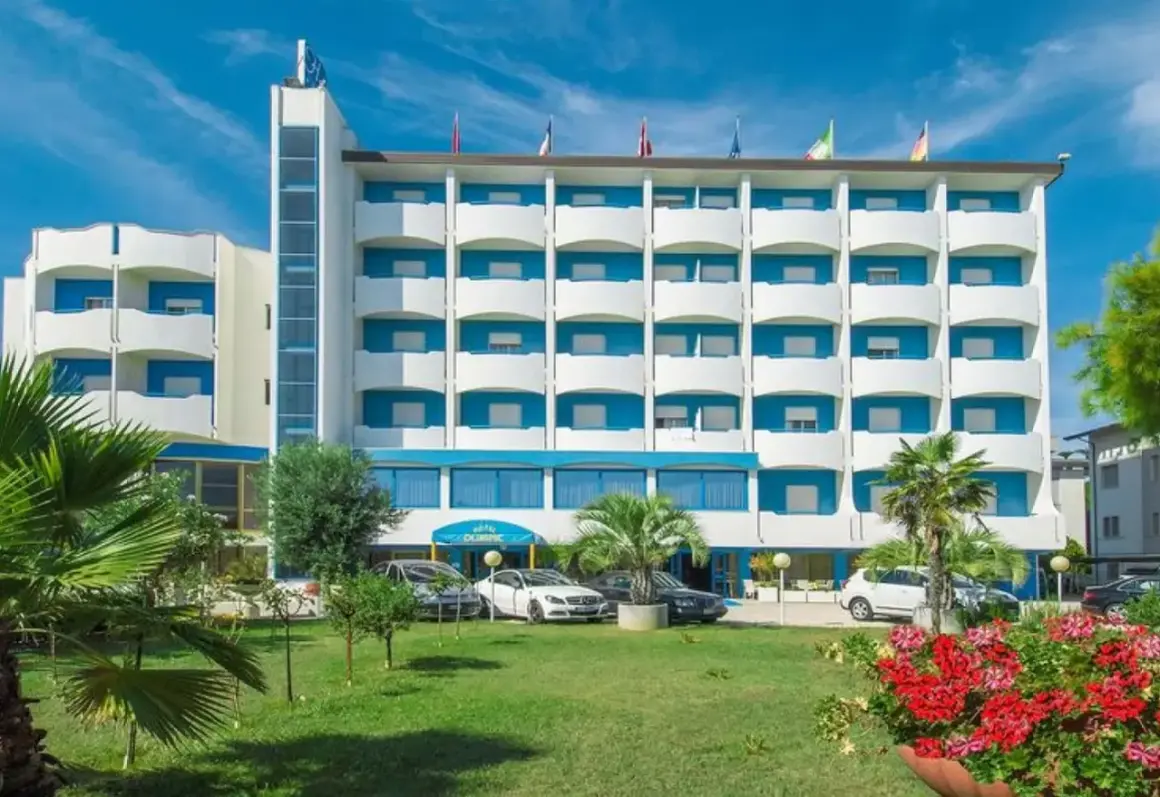 Hotel Olympic, un edificio bianco e blu con balconi, circondato da verde e fiori. Auto parcheggiate davanti.