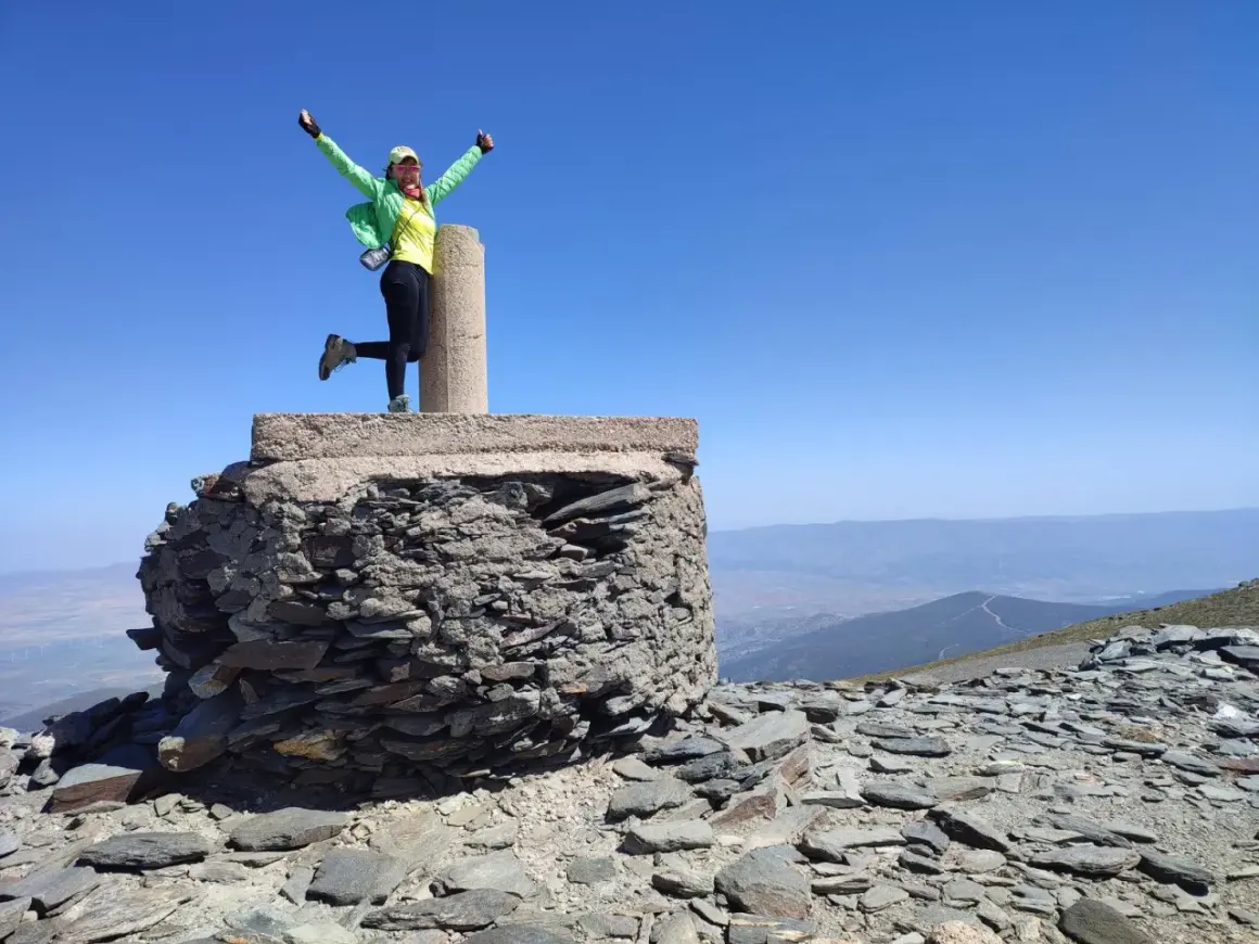 Mujer con el chullo puesto, brazos en alto, celebra en la cima de una montaña rocosa bajo un cielo azul despejado.