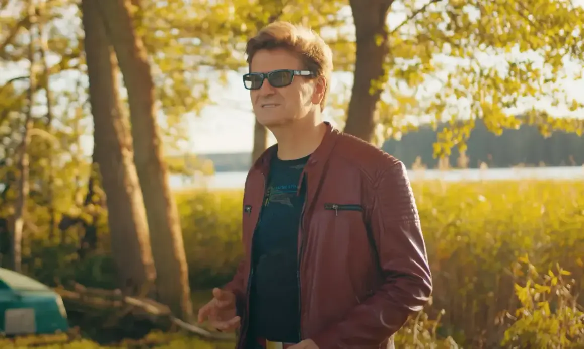 Mężczyzna w skórzanej kurtce, niczym król imperium disco polo, pozuje na tle jesiennego krajobrazu nad jeziorem.
