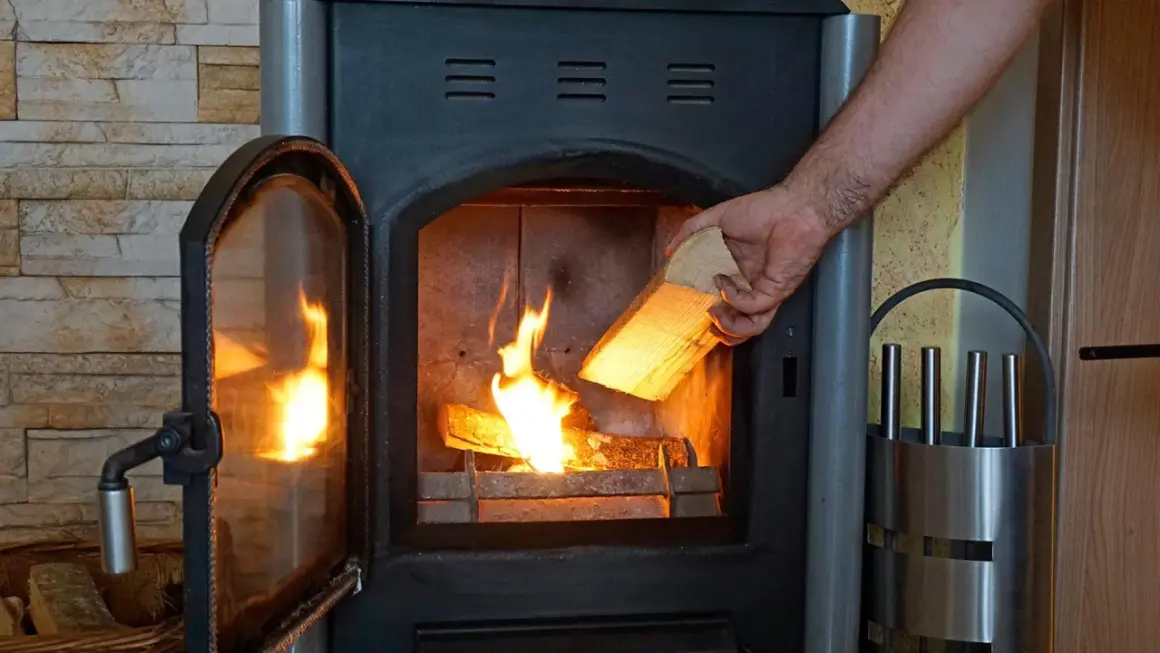 Hand legt Holzscheit in brennenden Kaminofen. Beachten Sie die Kaminofen Verordnung für sicheres Heizen.