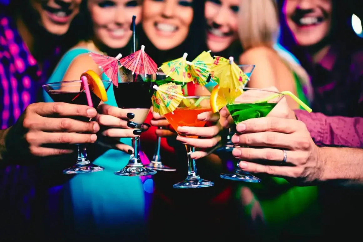 Amici brindano con cocktail colorati in un vivace night club a Monaco, tra risate e luci soffuse.