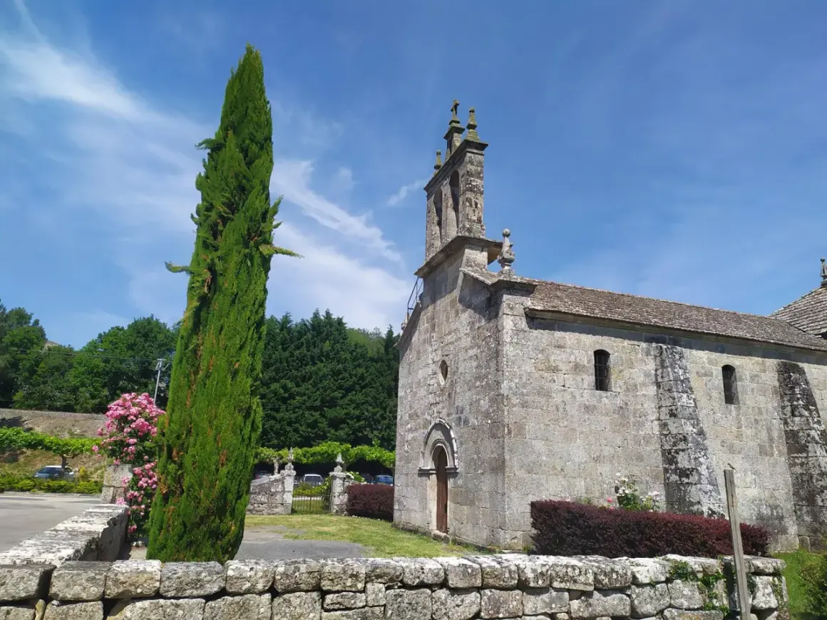 Iglesia de piedra bajo cielo azul, con un ciprés alto y flores rosas. Un lugar tranquilo en Cova da Moura.