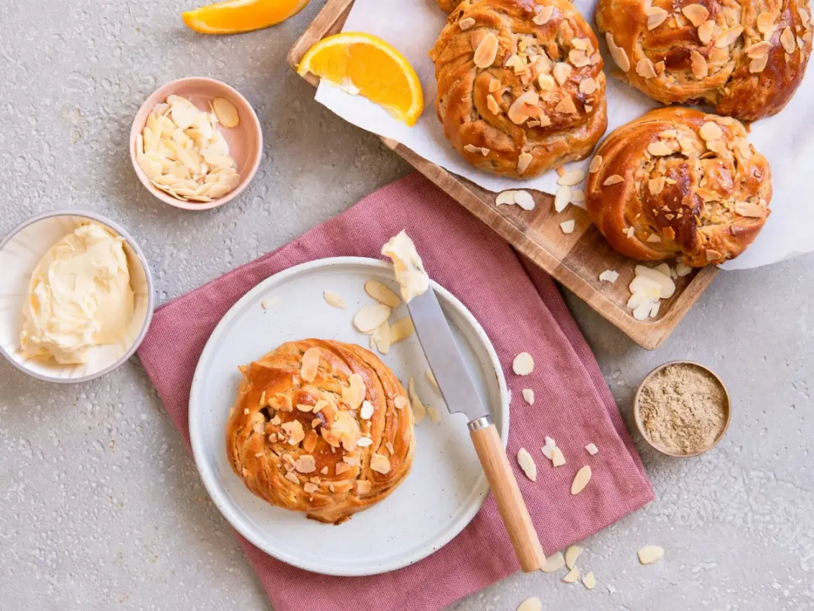 Goldbraune Kardamom-Knoten, bestreut mit Mandelblättchen, warten auf Butter und Orangenscheiben.