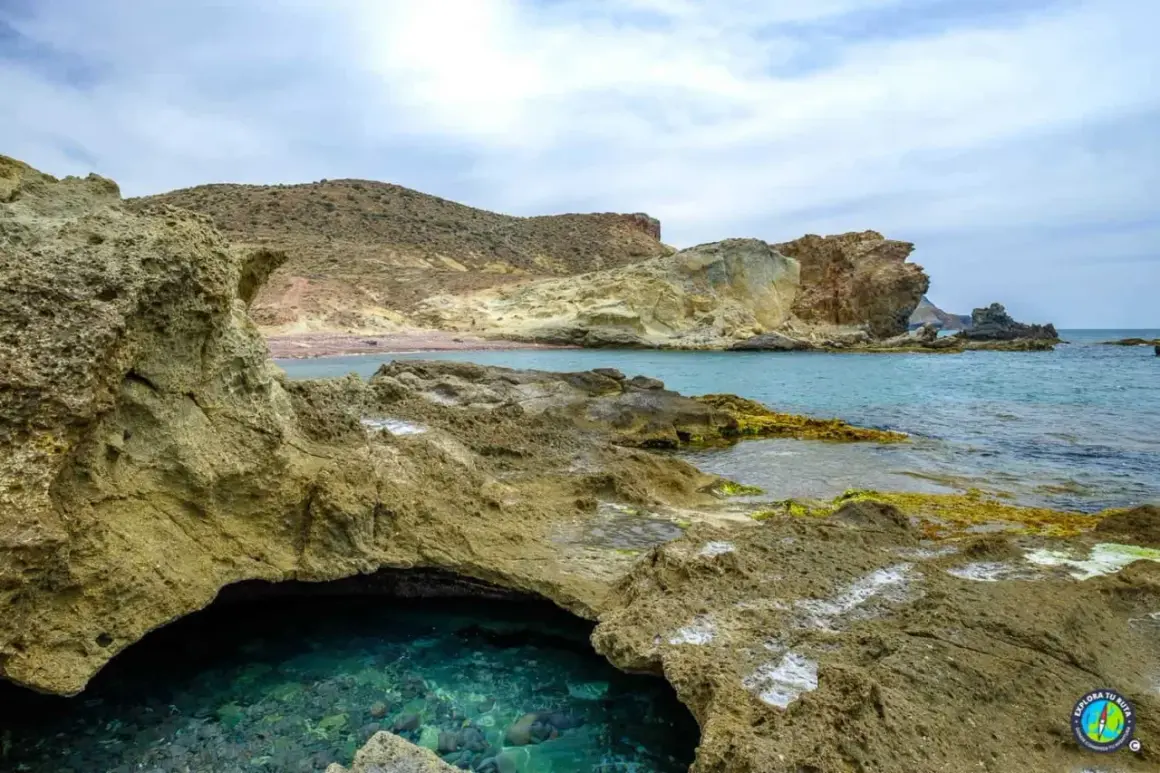 Agua turquesa en una cueva natural en la playa del carbón, con rocas volcánicas y montañas áridas al fondo.