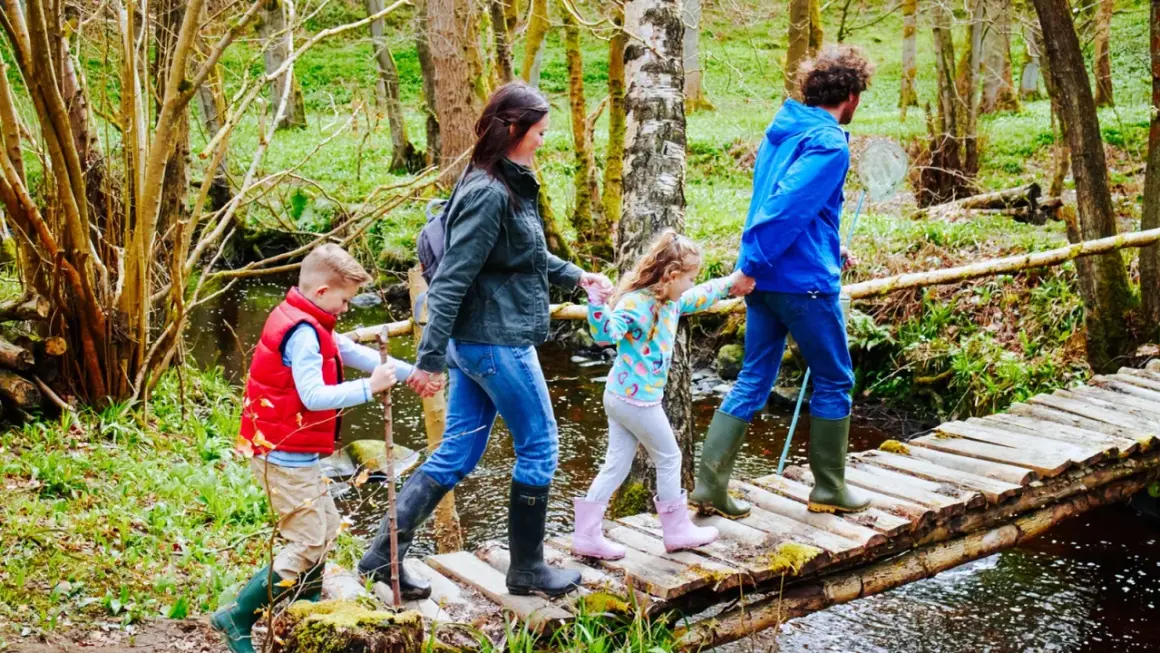 Familie überquert Holzbrücke auf einem erlebnispfad nrw. Vater hält Kescher, Kinder halten sich an Eltern.