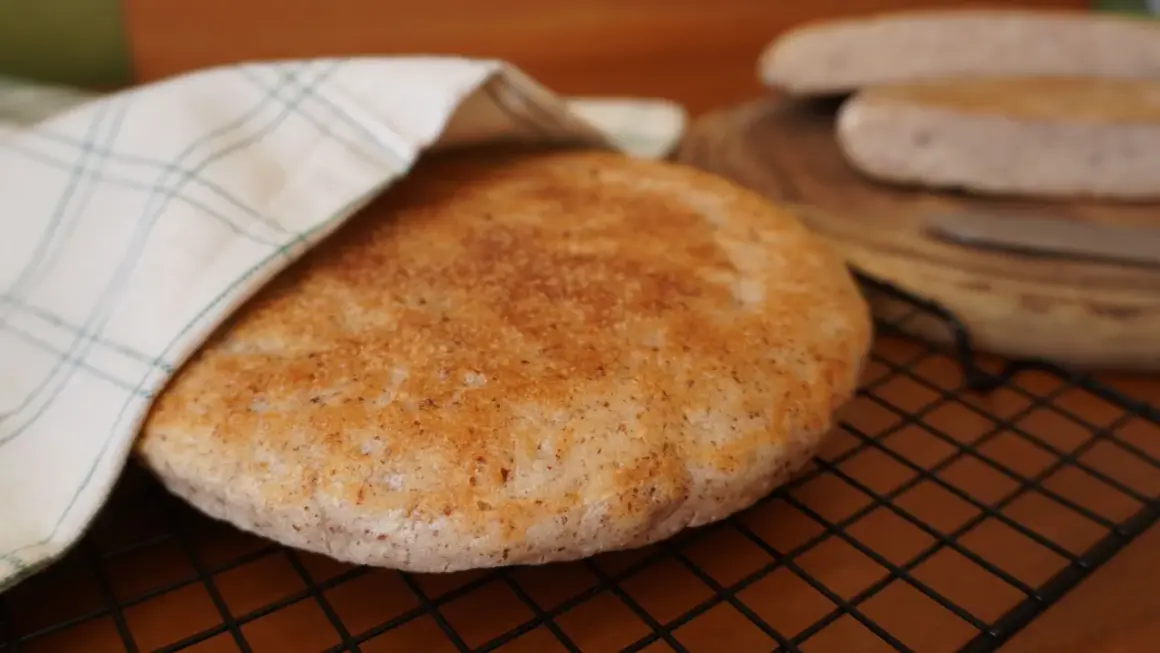 Pan de arroz redondo y dorado, cubierto parcialmente con un paño a cuadros, sobre una rejilla.