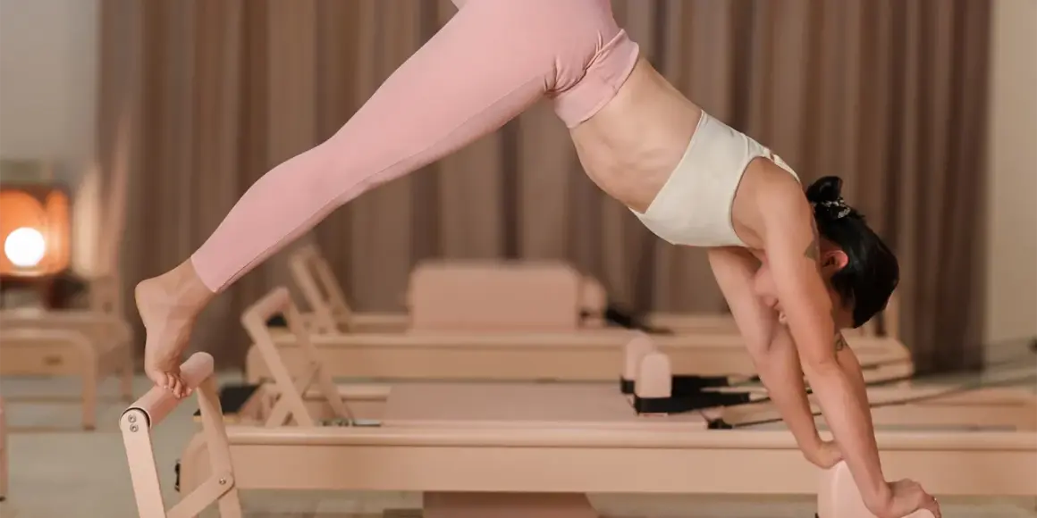 Kobieta w różowych legginsach wykonuje pozycję psa z głową w dół na reformerze. Pilates co daje? Wzmocnienie mięśni, elastyczność i harmonię.