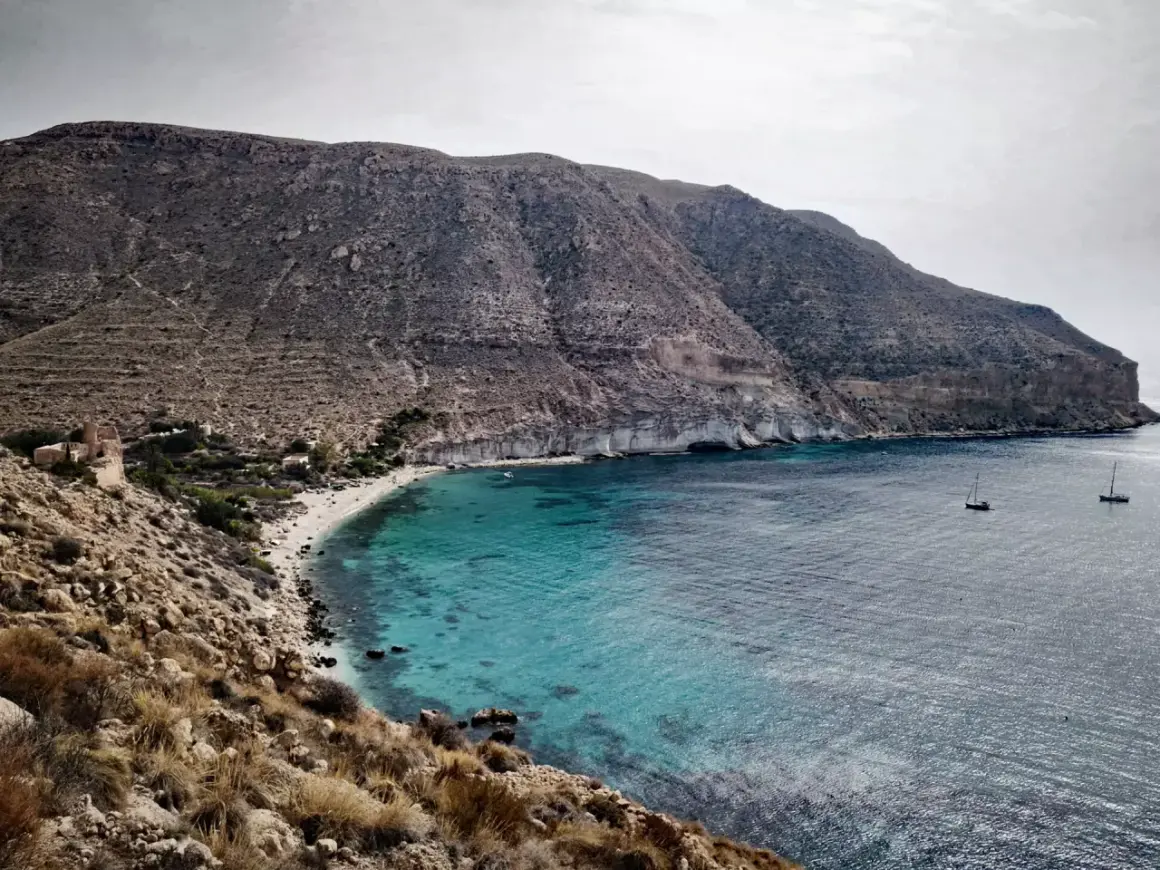 Bahía de aguas turquesas en la playa Cabo de Gata, con barcos navegando y montañas áridas al fondo.