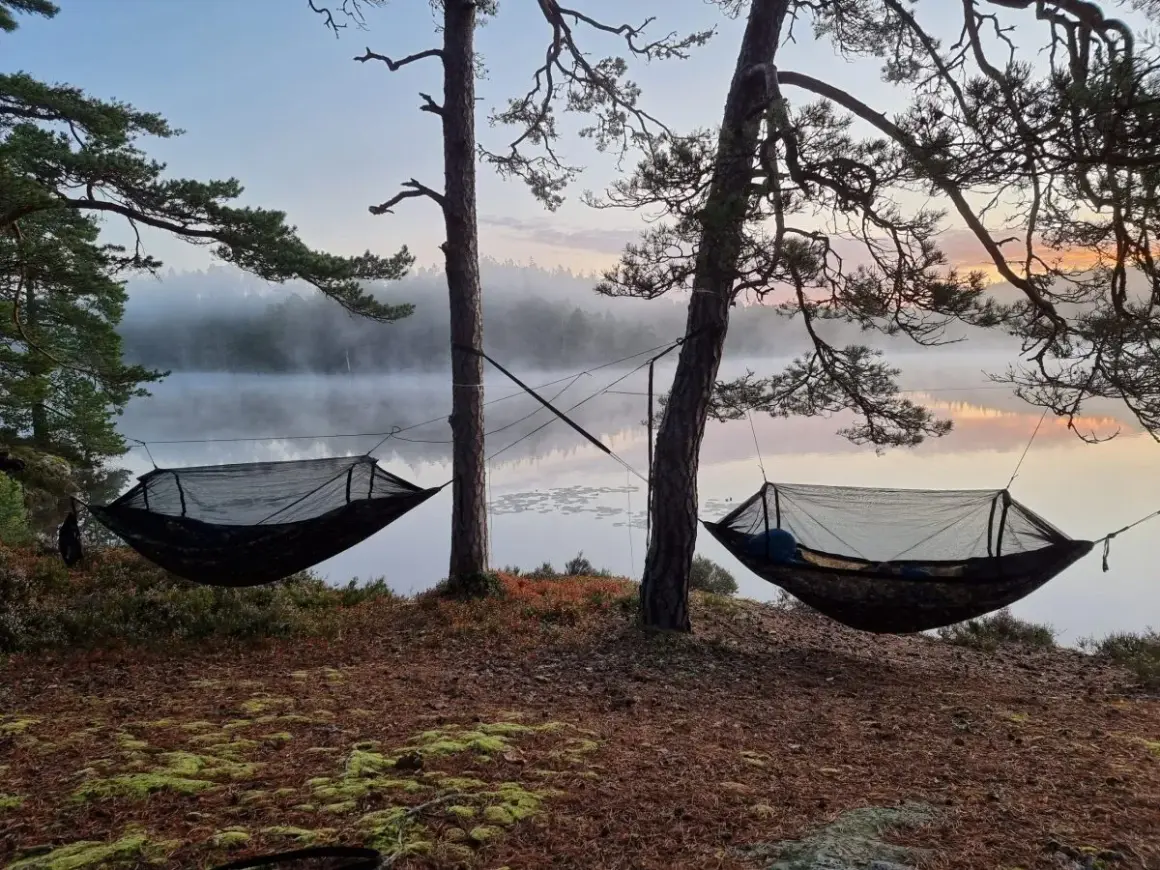 Två hängmattor redo för att sova i vid en dimmig sjö i gryningen. En person ligger redan och njuter av lugnet.