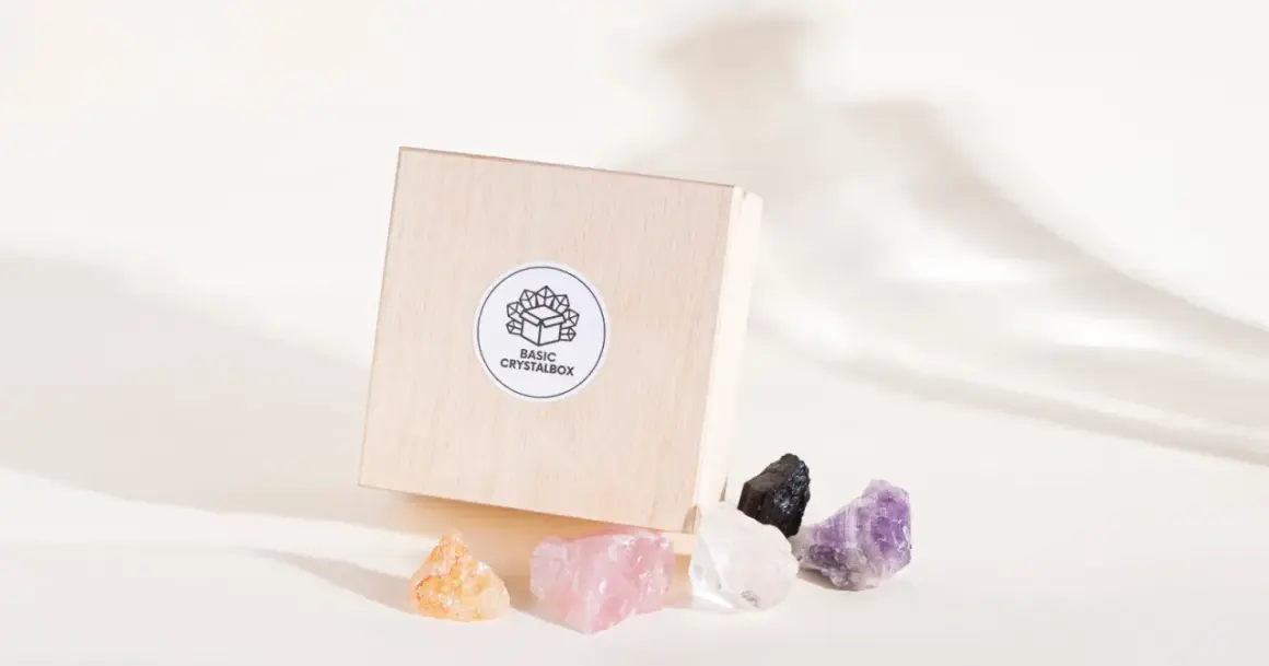 Drewniane pudełko z naklejką "BASIC CRYSTALBOX" i pięcioma surowymi kamieniami: bursztynowym, różowym kwarcem, kryształem górskim, czarnym turmalinem i ametystem. Każdy cenny kamień emanuje naturalnym blaskiem.