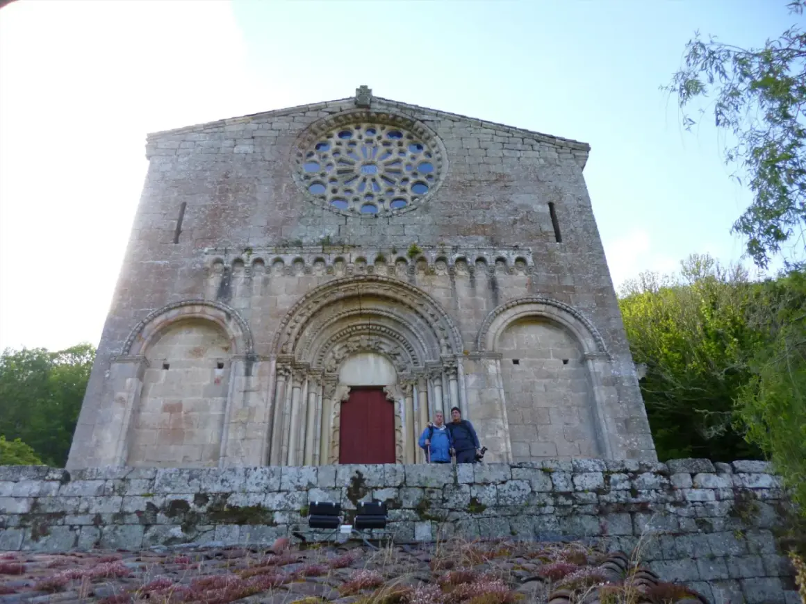 Dos personas posan frente a la fachada románica de Santo Estevo de Ribas de Miño, con su rosetón y portal detallados.
