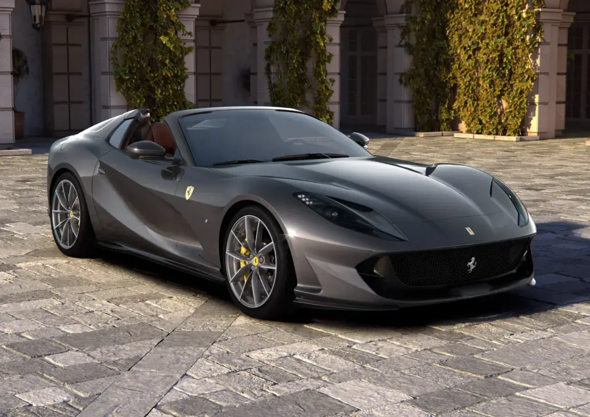 Szary kabriolet Ferrari 812 GTS z żółtymi zaciskami hamulcowymi stoi na brukowanym dziedzińcu przed budynkiem porośniętym bluszczem.