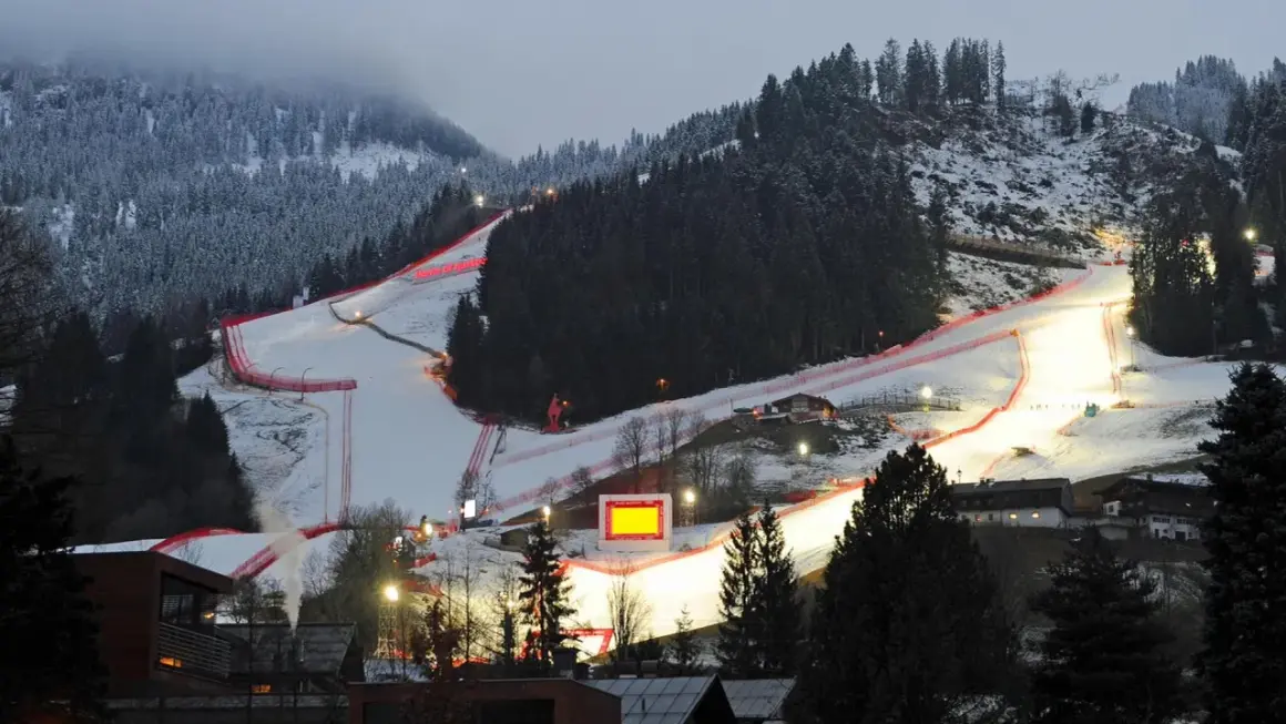 Die Skirennstrecke, beleuchtet für die Nacht, mit roten Banden und einem roten Hahnkamm-Symbol.