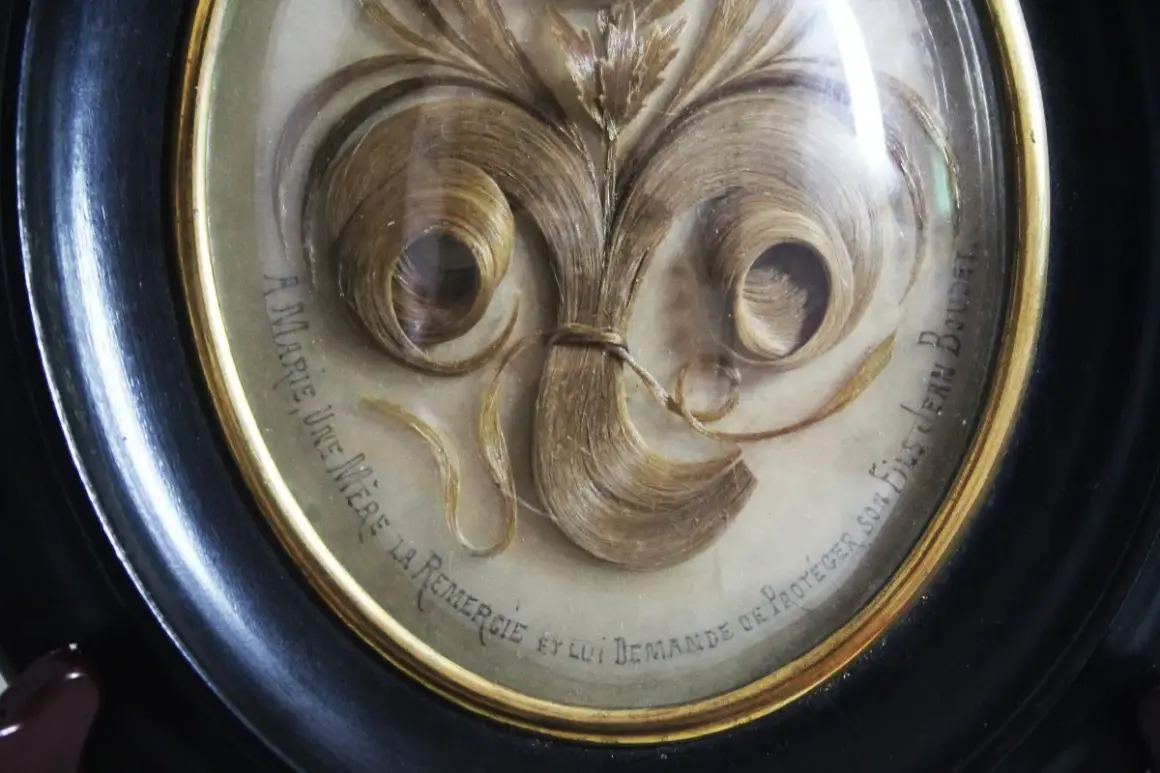 Intricate hair art in a framed oval, featuring swirling locks of blonde hair arranged like a floral bouquet. Text reads "A Marie, une mère la remercie et lui demande de protéger son fils Jean Boudet.