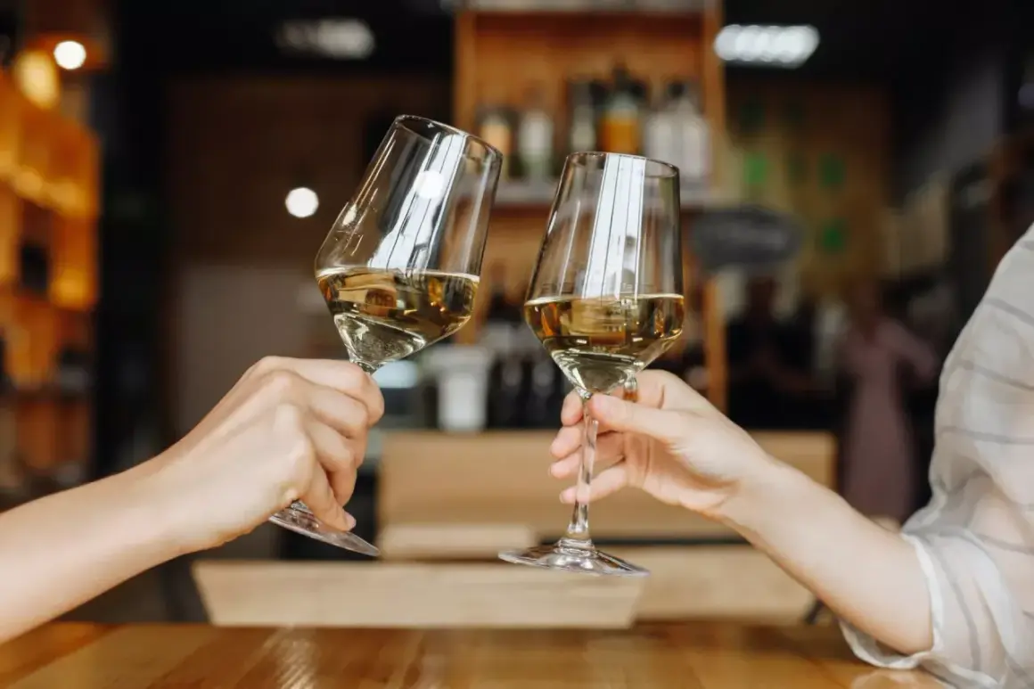 Zwei Hände halten Gläser mit einem goldenen Cuvée Wein. Ein Toast auf den Genuss in gemütlicher Atmosphäre.
