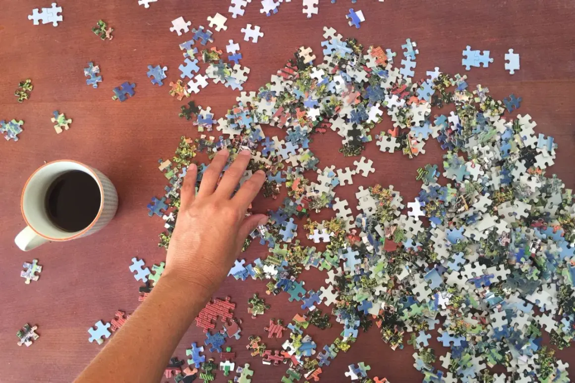 Ręka układa puzzle do układania na drewnianym stole obok kubka kawy.