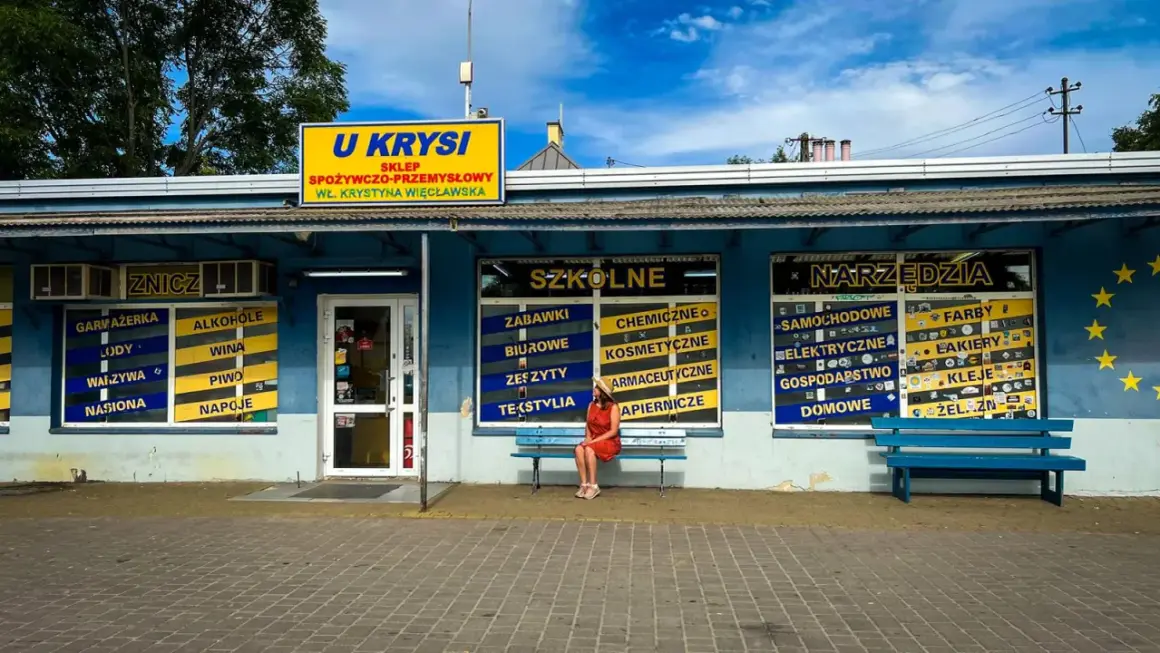 Dziewczyna w czerwonej sukience siedzi na ławce przed sklepem "U Krysi", gdzie można kupić wszystko, od zniczy po narzędzia.