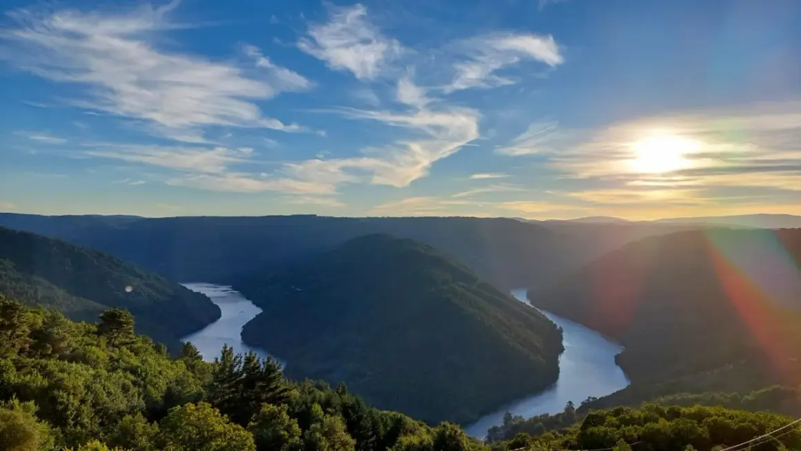 Un río serpentea entre montañas verdes, bajo un cielo azul con nubes finas. El sol se pone, tiñendo el horizonte de dorado. Parece el cabo do mundo.