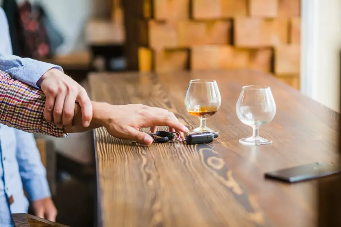 Ręka powstrzymuje drugą przed sięgnięciem po kluczyki do samochodu. Na stole stoją kieliszki z alkoholem, sugerując ryzyko utraty prawa jazdy za alkohol.