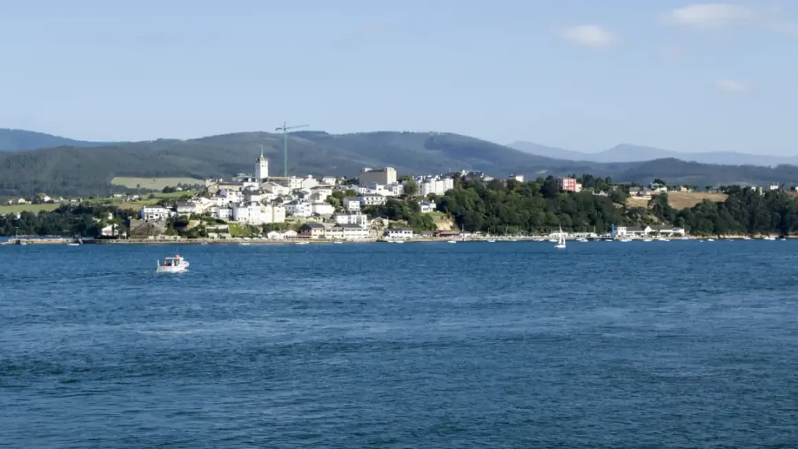 Vista del mirador de Ribadeo con casas blancas y el mar azul. Un pequeño barco navega en la distancia.