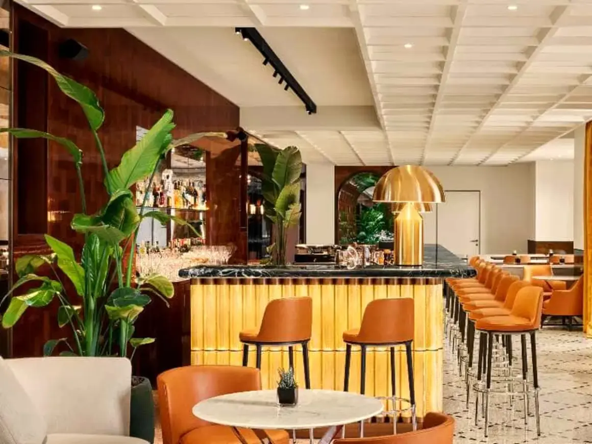 Interni eleganti del Bulgari Hotel Roma: bancone bar dorato, sedie in pelle arancione, piante lussureggianti e un'atmosfera sofisticata.