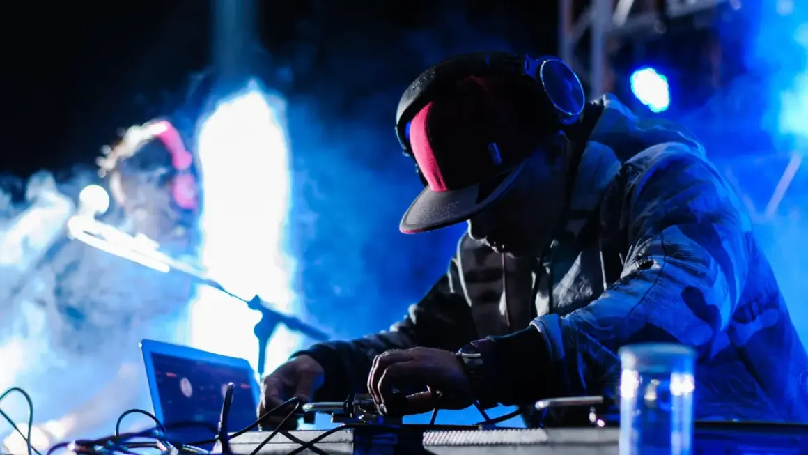 Un techno dj con cuffie e cappellino rosso mixa musica su un palco illuminato da luci blu e fumo.