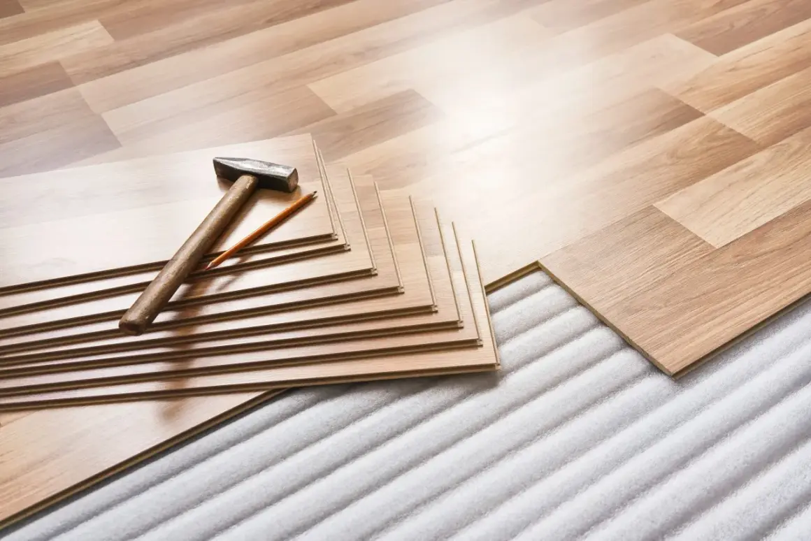 Tablas de suelo laminado apiladas con martillo y lápiz, listas para instalar sobre una base que protege de la humedad que sube del suelo.