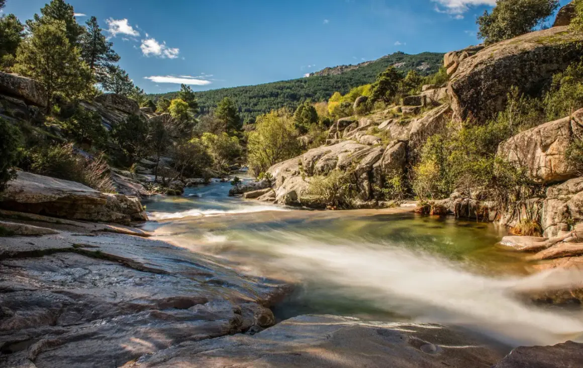 Río serpenteante entre rocas y vegetación, con una montaña boscosa al fondo, un paisaje idílico cerca de Madrid.