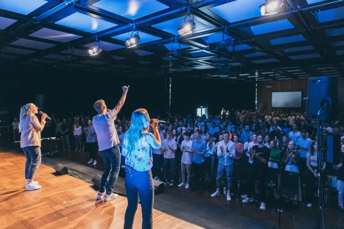 Junge Leute singen auf einer Bühne vor einer großen Menge. Ein Mann hebt die Hand, als ob er eine Botschaft verkündet. Eine moderne Kirche, die urbanes Leben und Gemeinschaft vereint.