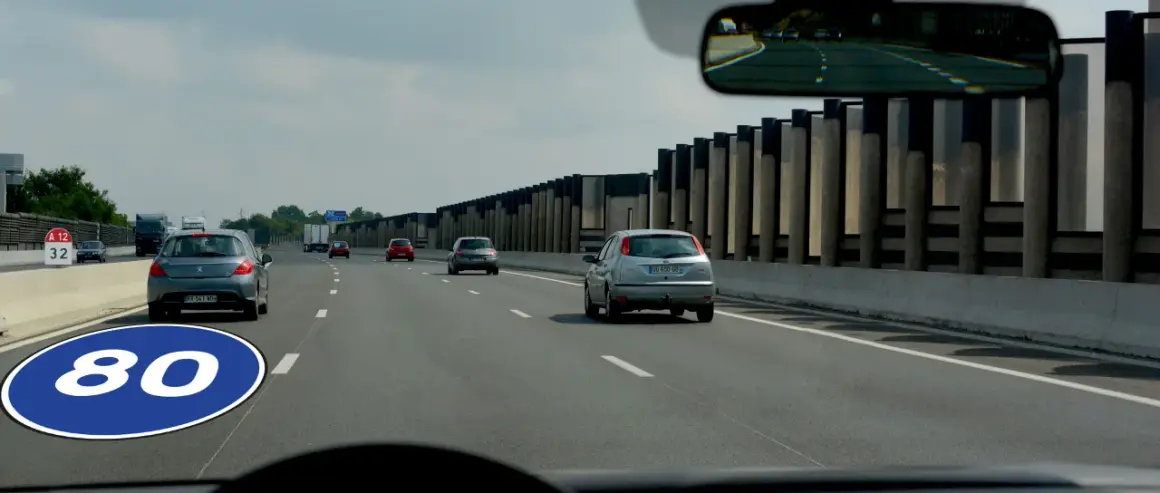 Panneau de signalisation au sol indiquant la vitesse minimale autoroute de 80 km/h. Plusieurs voitures circulent sur la voie rapide.