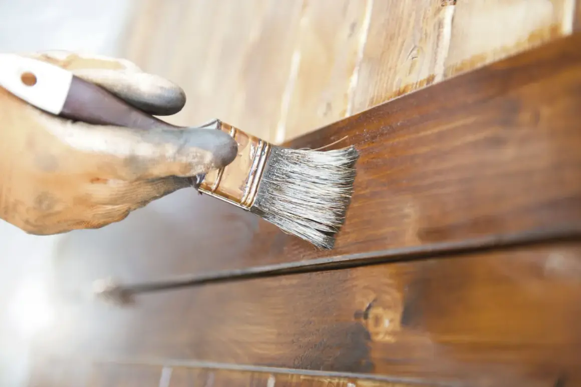 Main gauche gantée appliquant du vernis sur des lattes de bois brun pour vernir un meuble.
