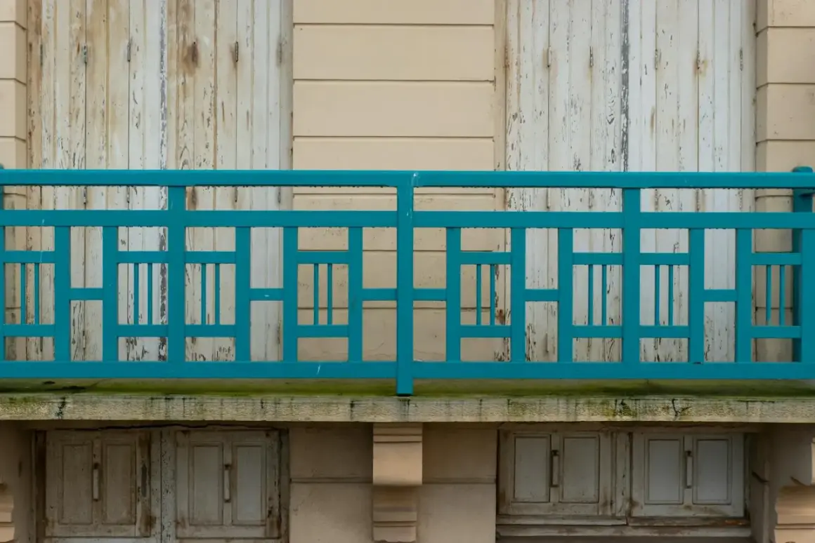 Turkusowe, geometryczne mocowanie balustrady od czoła, z widocznymi odrapanymi drzwiami poniżej.