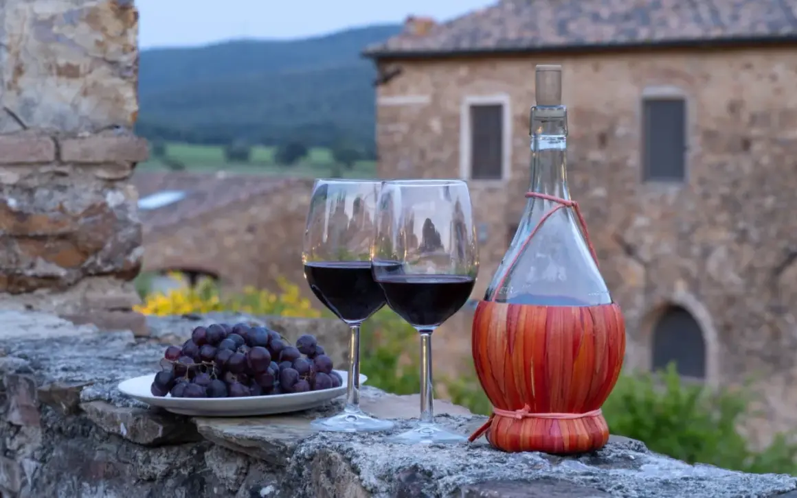 Dwa kieliszki czerwonego wina, kiść winogron i butelka w koszyczku, na tle kamiennego muru i krajobrazu regionu Chianti.